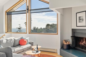A modern living room with a fireplace, a sofa with cushions, a coffee table, and a large window with a view. - Hawk Haven | Ocean Views, Hot Tub & Cozy Fireplace (Sea Ranch)