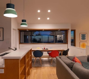 A modern kitchen with a dining area, a sofa, and a window with a view of trees. - Hawk Haven | Ocean Views, Hot Tub & Cozy Fireplace (Sea Ranch)