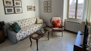 Un salon comprenant un canapé, un fauteuil, une table basse et un téléviseur. Des tableaux encadrés ornent le mur, et une fenêtre offre une vue sur les bâtiments environnants.