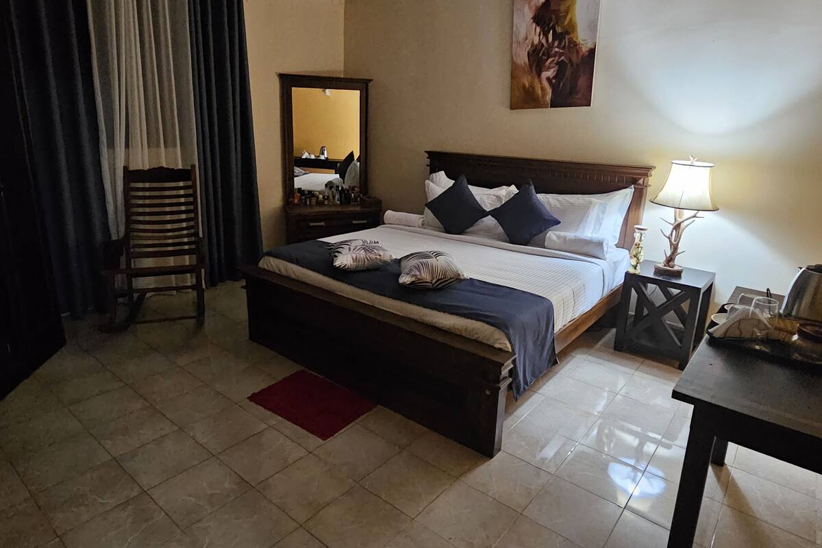 A hotel room with a bed, a nightstand, a lamp, a chair, a mirror, and a painting on the wall.