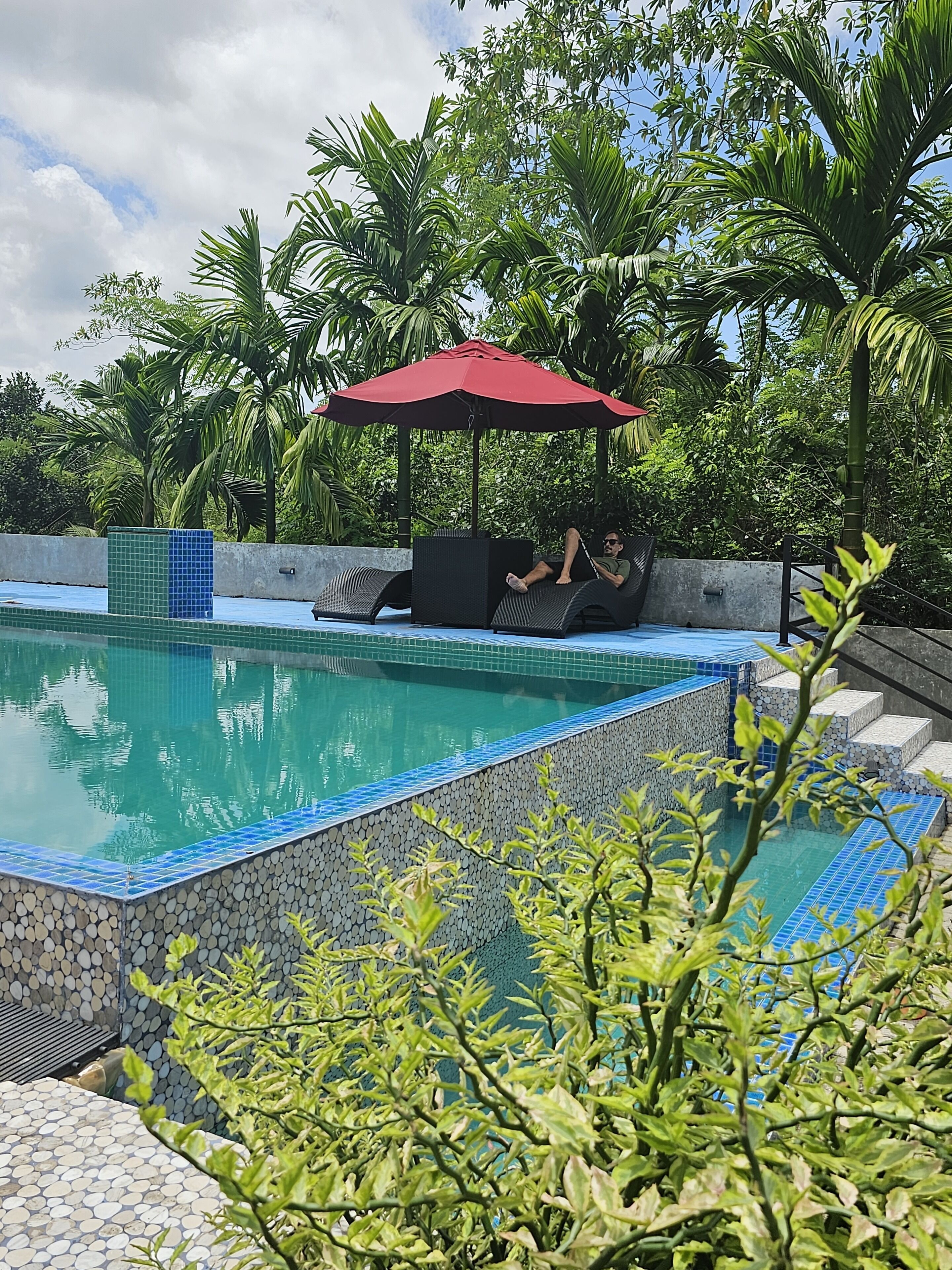 A swimming pool with tiled edges, surrounded by lush greenery and a red umbrella.