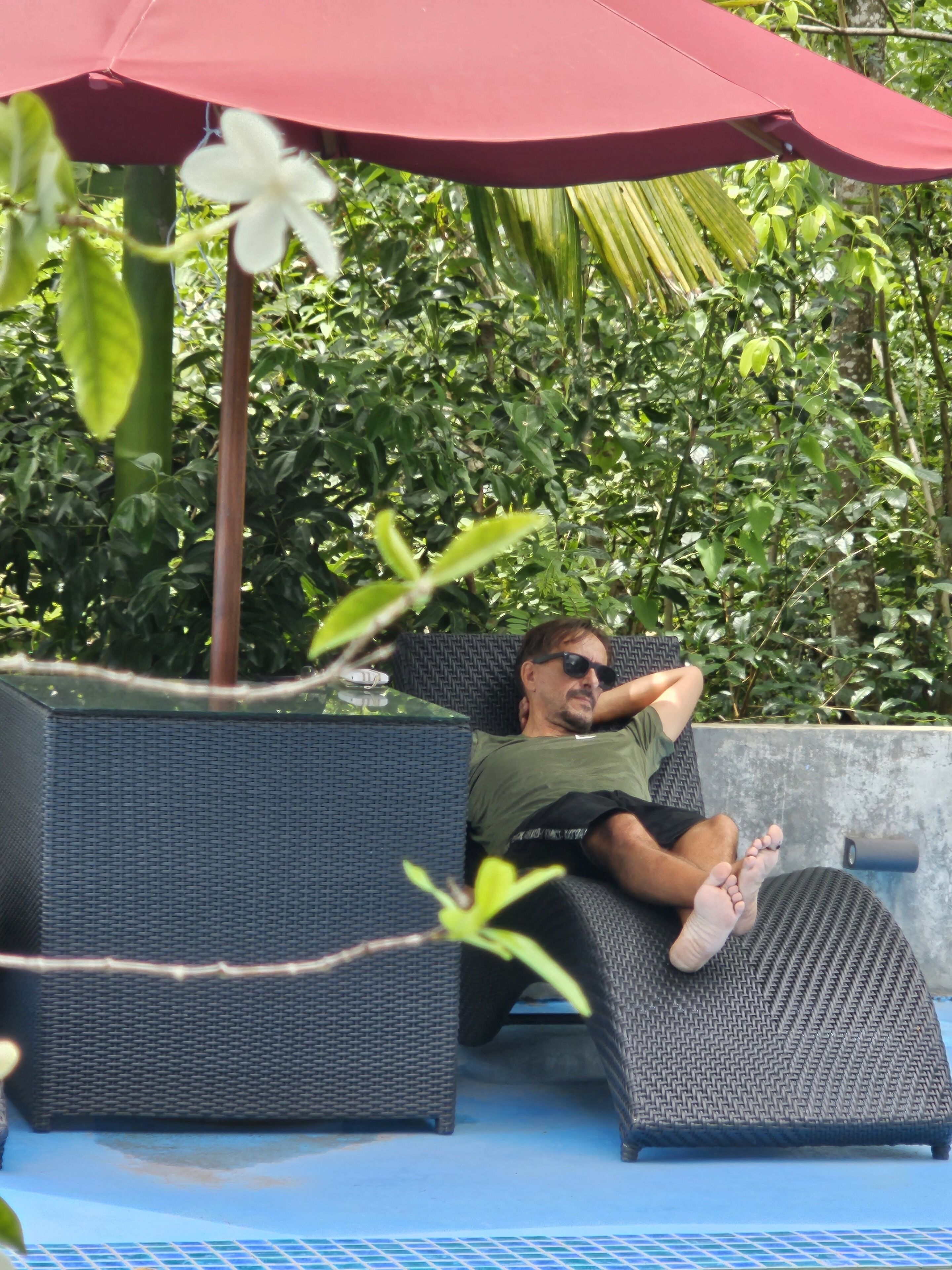 Un homme se détend sur un fauteuil en osier, à l'ombre d'un parasol rouge, entouré d'une végétation luxuriante.