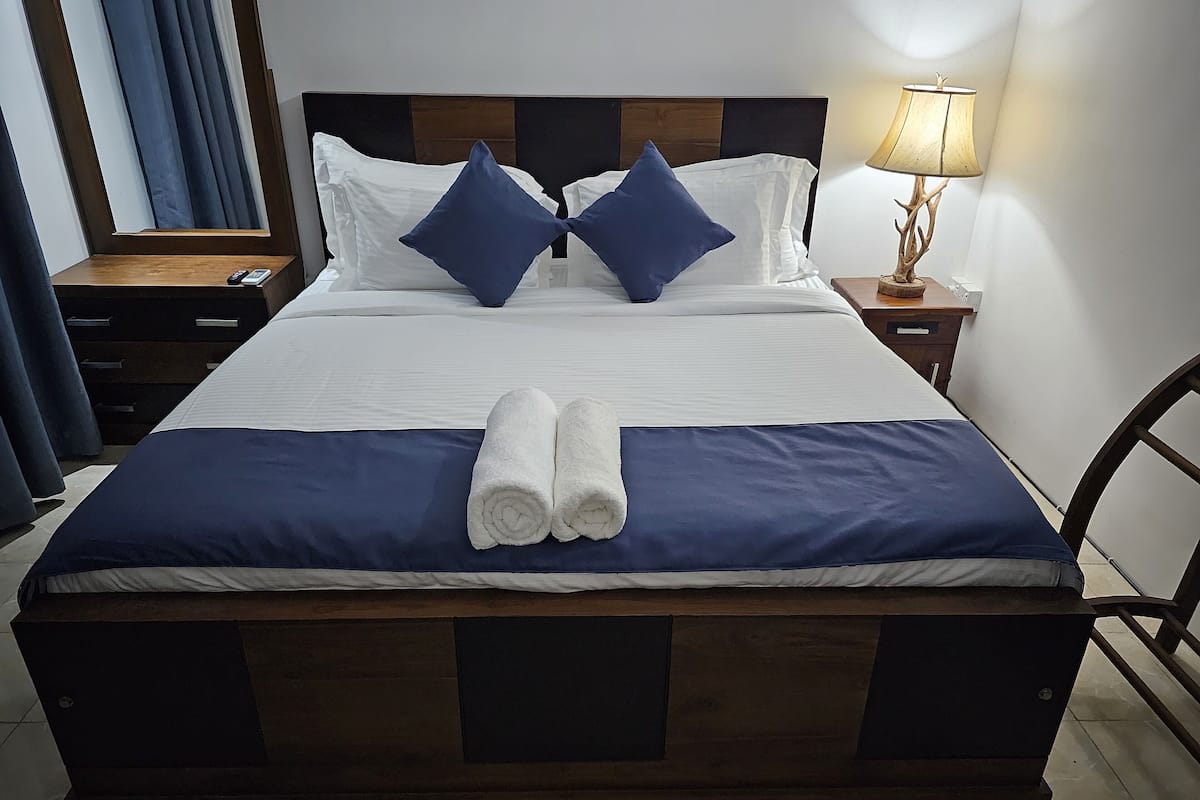 A neatly made bed with blue and white bedding, two rolled towels, a wooden headboard, a bedside lamp, and a nightstand with a phone.