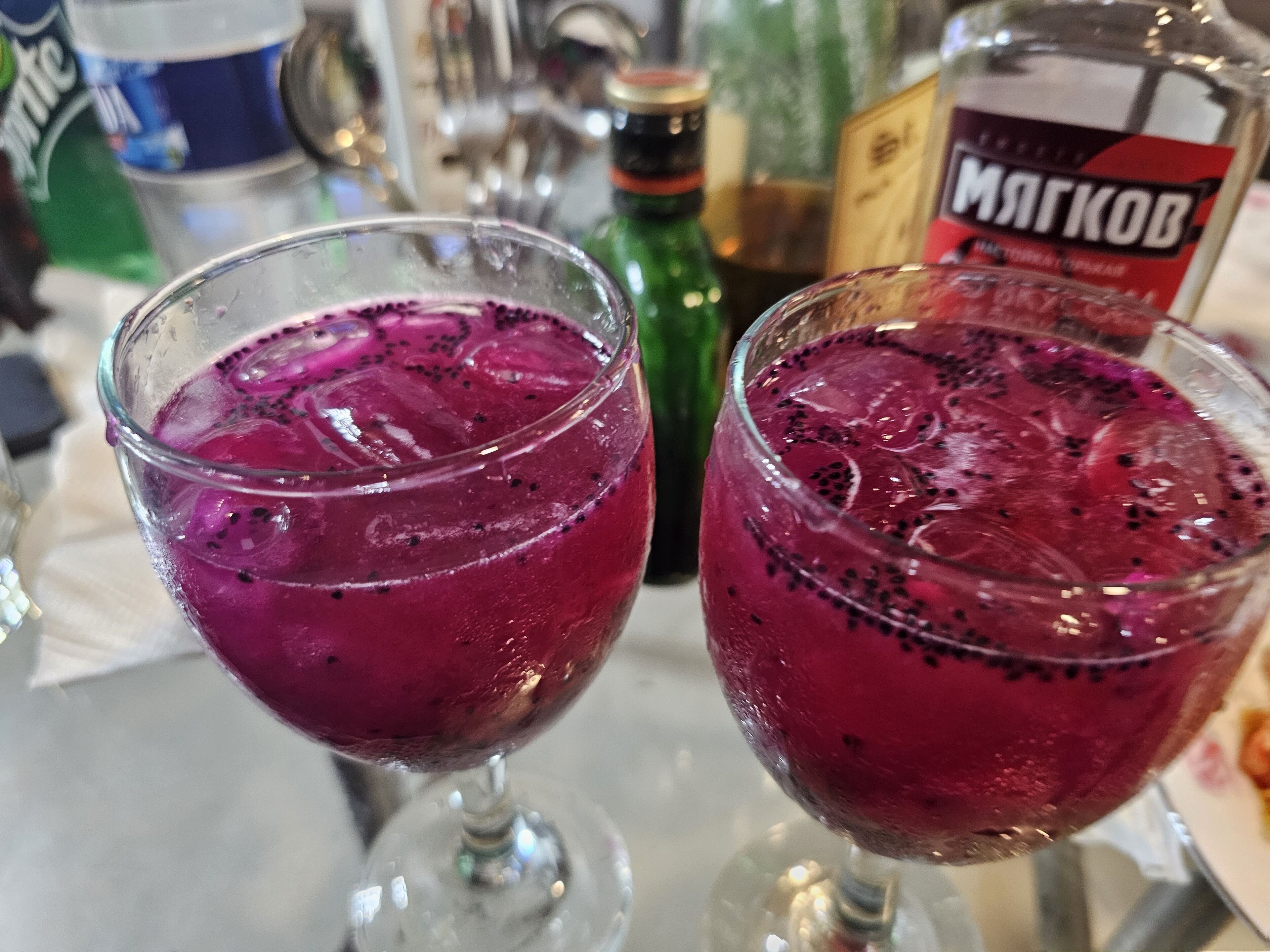 Deux verres contenant une boisson violette, avec des glaçons et de petites taches noires, peut-être des graines, sont posés sur une table où se trouvent différentes bouteilles, et une bouteille verte est visible à l’arrière-plan.