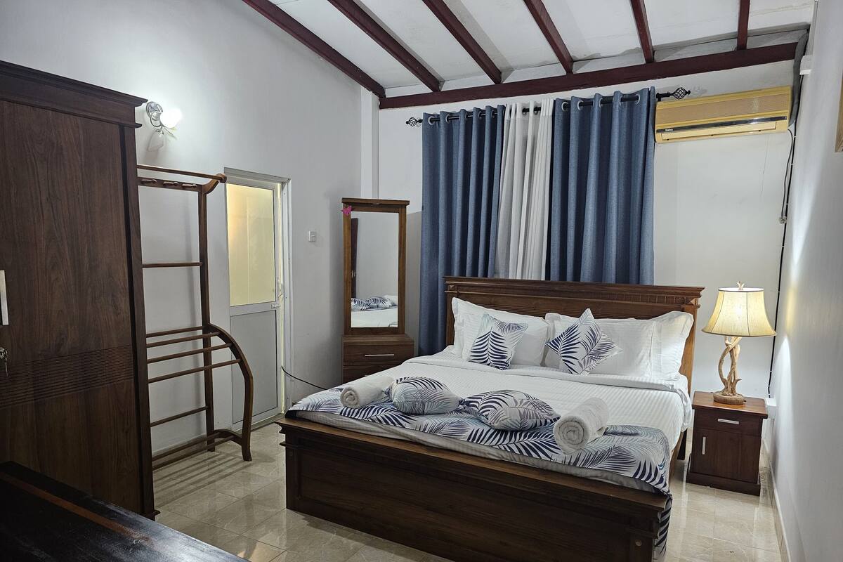 A bedroom with a wooden bed, a nightstand with a lamp, a dresser, a mirror, and a window with curtains.