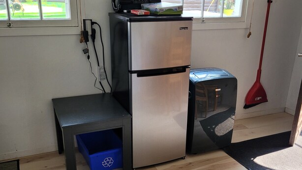 A compact kitchen with a refrigerator, a small table, and a blue recycling bin. - The Cottages at Gull & Castle Cabin #1 (Wasagaming)