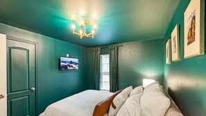 A bedroom with teal walls, a bed with white linens, a mounted TV, and a chandelier.