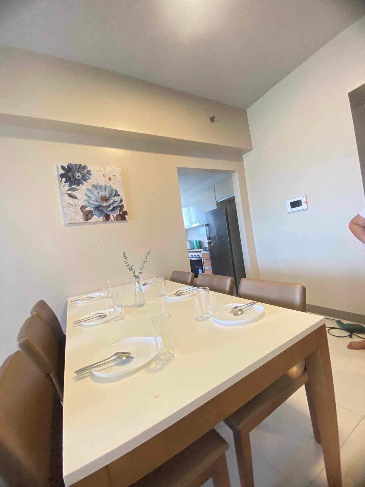 A dining area with a white table set for four, surrounded by wooden chairs. A decorative floral painting is on the wall above the table.