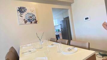 A dining area with a white table set for four, surrounded by wooden chairs. A decorative floral painting is on the wall above the table.