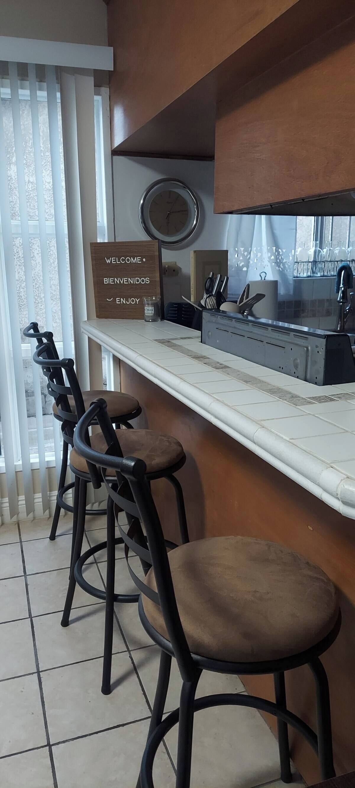 A kitchen area with a bar counter, stools, a welcome sign, and a clock on the wall.