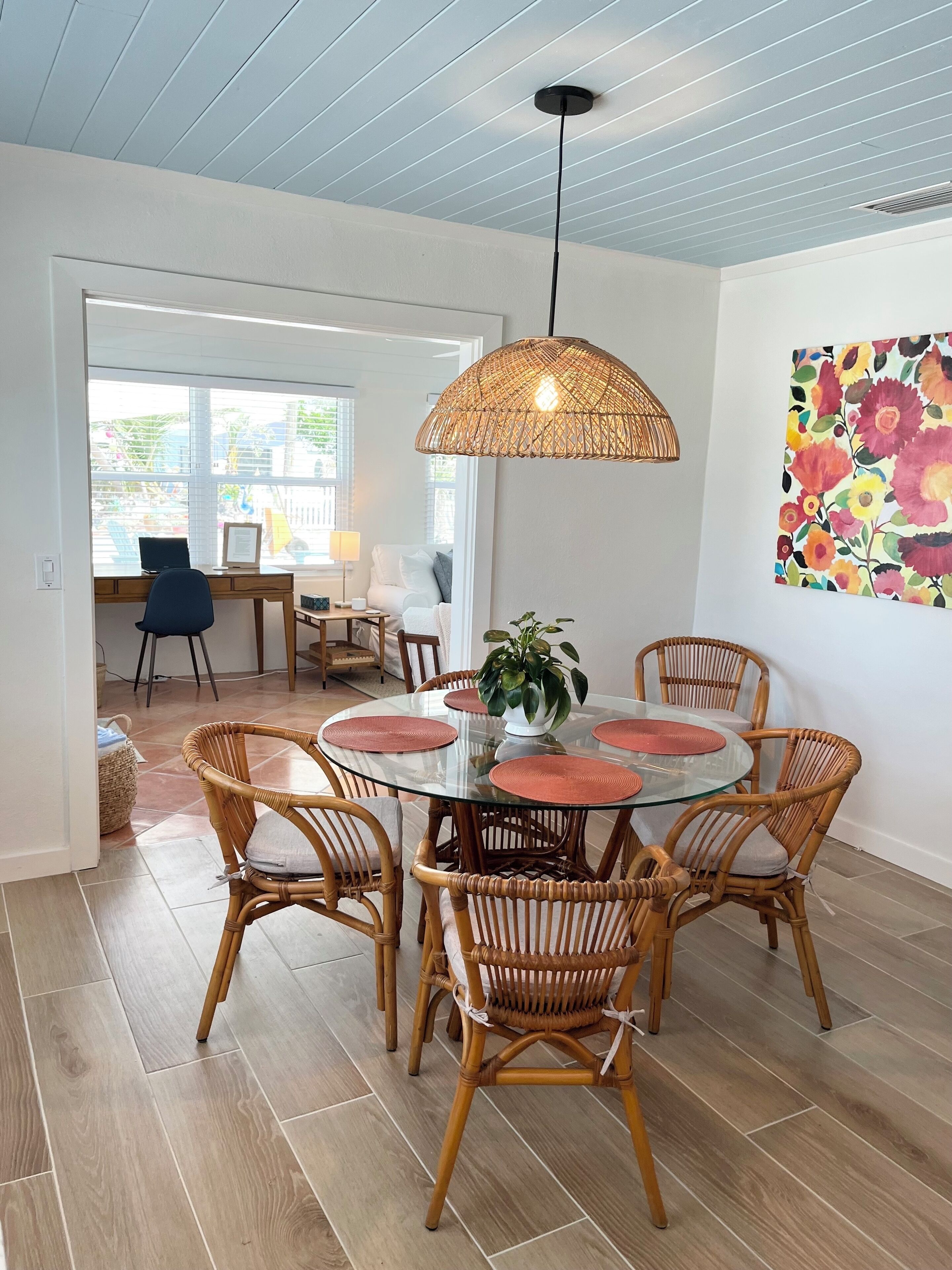Un coin repas avec une table ronde en verre entourée de chaises en osier, un luminaire suspendu en fibres tressées et une œuvre d’art murale aux motifs floraux et aux couleurs vives.