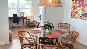 A dining area with a round glass table surrounded by wicker chairs, a woven pendant light, and a colorful floral wall art piece. - FLASH SALE - just steps to beach. 2BR/1BA.Dogs welcome! Perfect ratings. (Englewood)