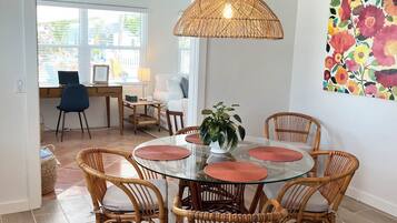 Un coin repas avec une table ronde en verre entourée de chaises en osier, un luminaire suspendu en fibres tressées et une œuvre d’art murale aux motifs floraux et aux couleurs vives.