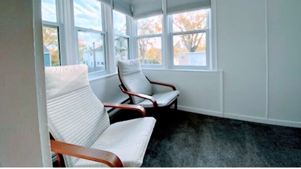 A room with two white upholstered chairs, a dark carpet, and large windows with a view of trees and buildings. - Retraite Saint Boniface/St. Boniface Retreat (Winnipeg)