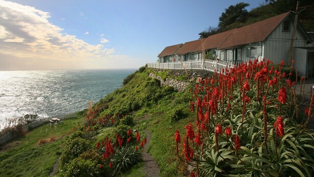 A coastal building with a white fence, red flowers, and a view of the ocean. - Lucia Lodge (Big Sur)
