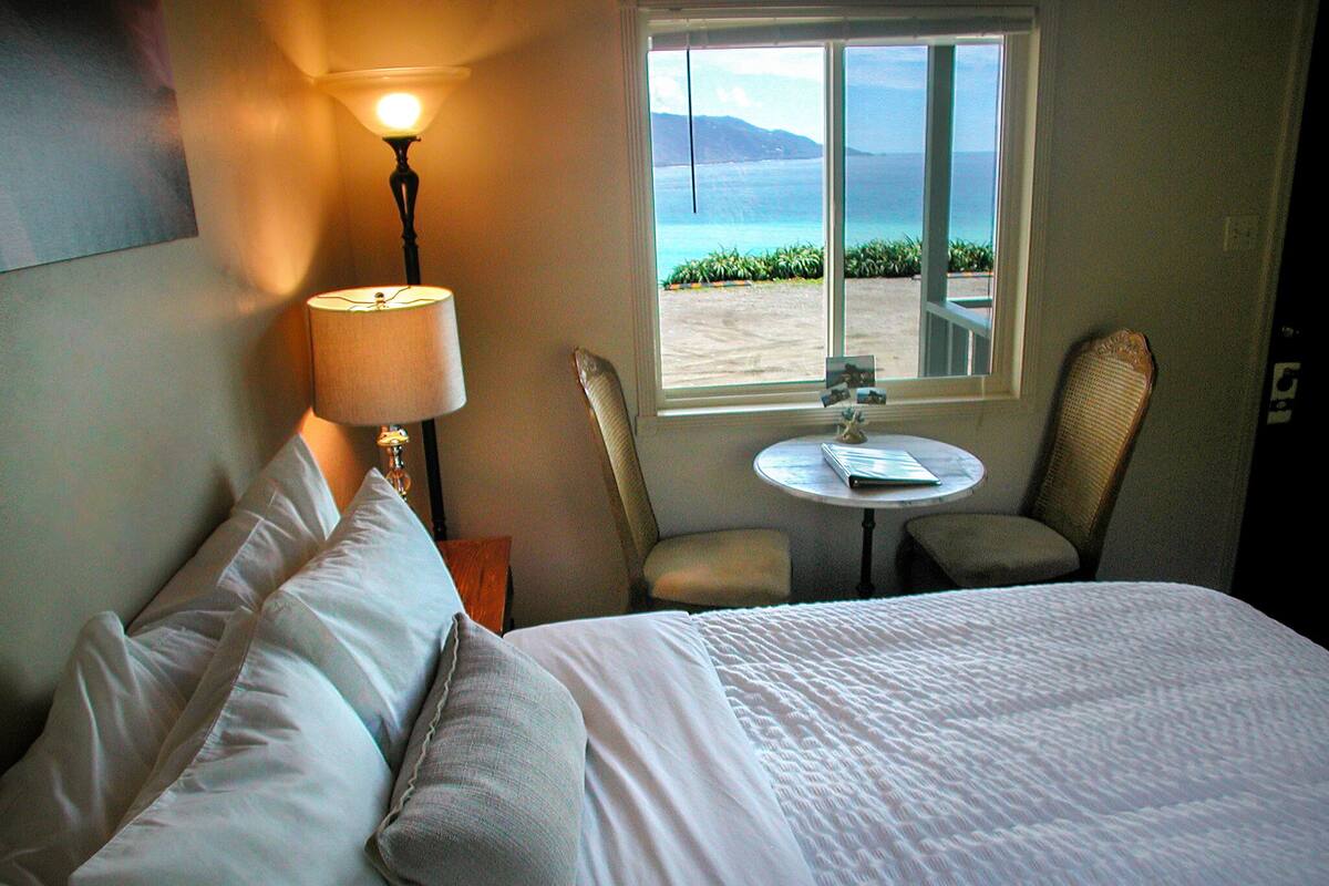 A hotel room with a bed, a lamp, a small table, two chairs, and a window with a view of the sea.