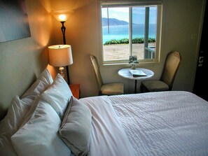 Une chambre d’hôtel avec un lit, une lampe, une petite table, deux chaises et une fenêtre donnant sur la mer.