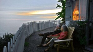 Two people sitting on a wooden bench overlooking a body of water, with a white fence and a building with a lit window in the background. - Lucia Lodge (Big Sur)