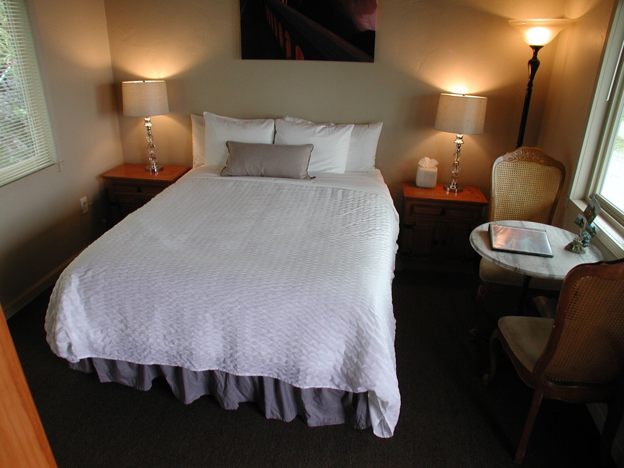 A neatly made bed with white linens, two bedside tables with lamps, a chair, and a small table with a laptop.