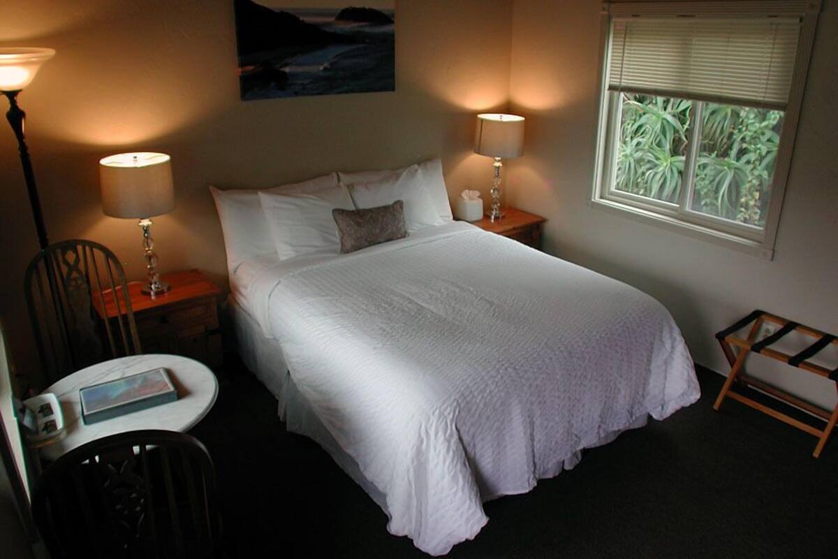 A neatly arranged bedroom with a large bed, two bedside lamps, a small table with a book, a chair, and a window with blinds.