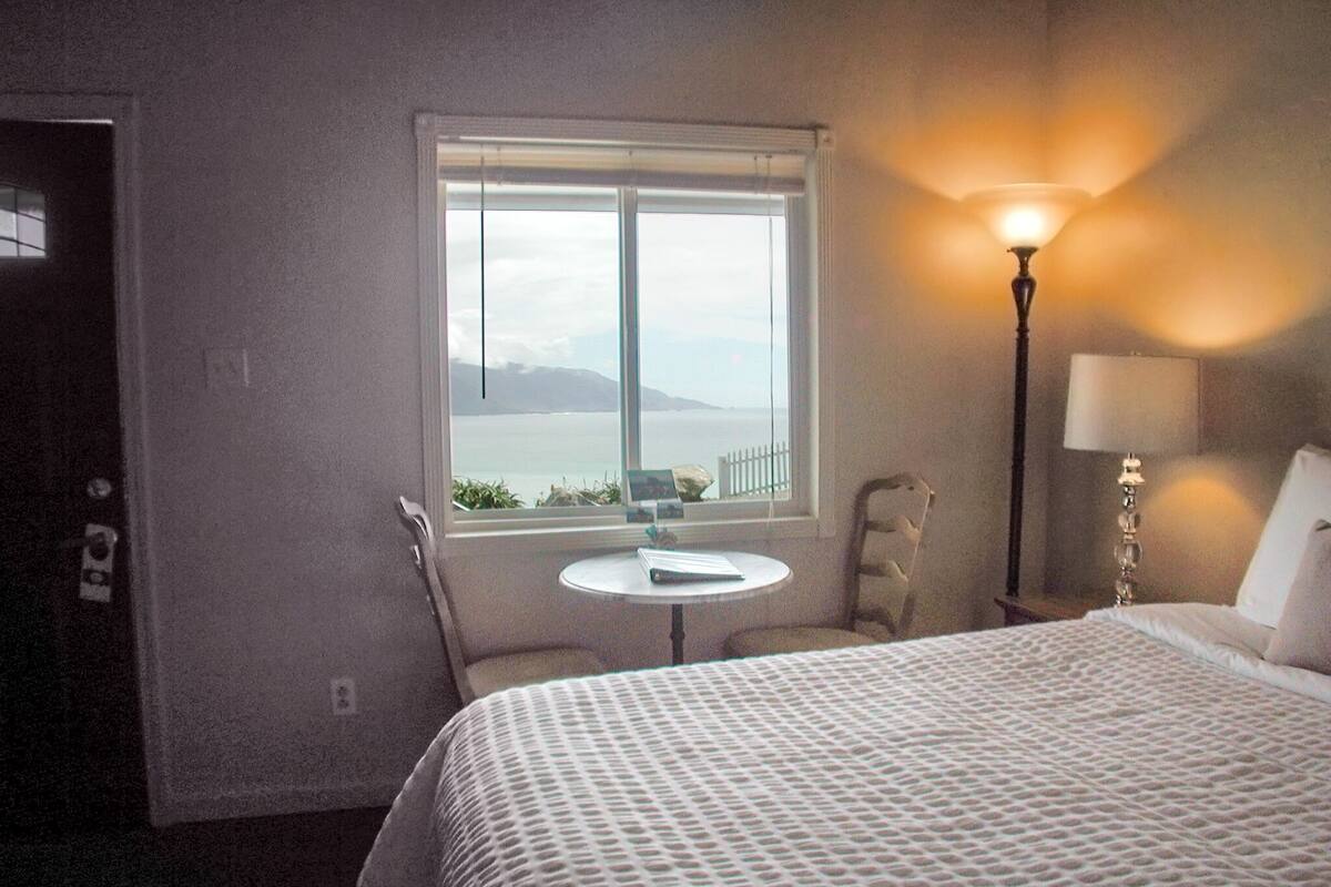 A hotel room with a bed, a small round table, a chair, a lamp, and a window with a view of the sea and mountains.