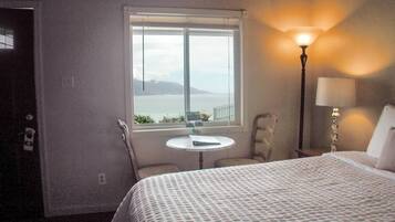 A hotel room with a bed, a small round table, a chair, a lamp, and a window with a view of the sea and mountains.
