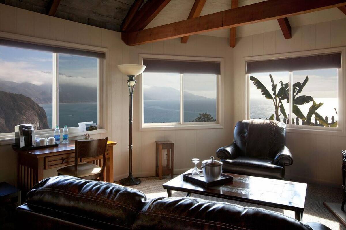 A cozy living room with a view of the ocean, a wooden desk with a lamp, a leather armchair, and a coffee table.