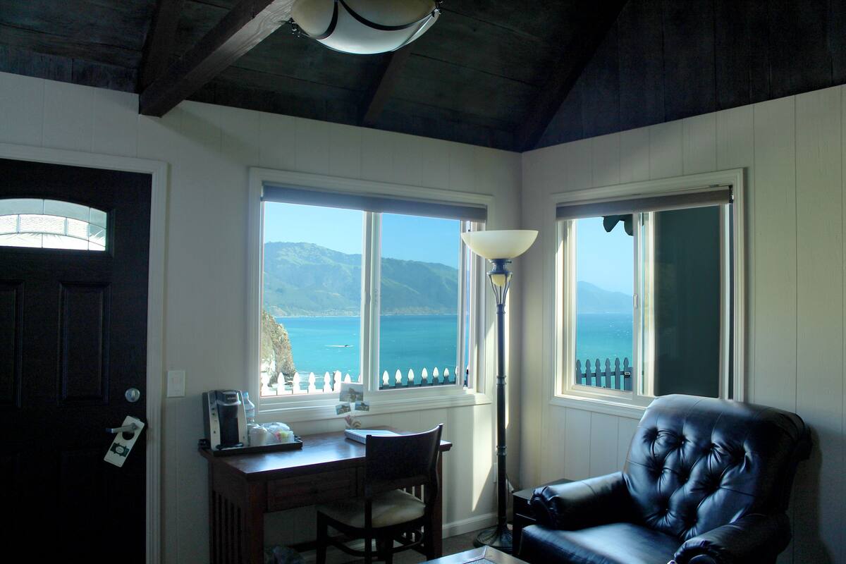 A room with a desk, chair, and a view of the sea through large windows.