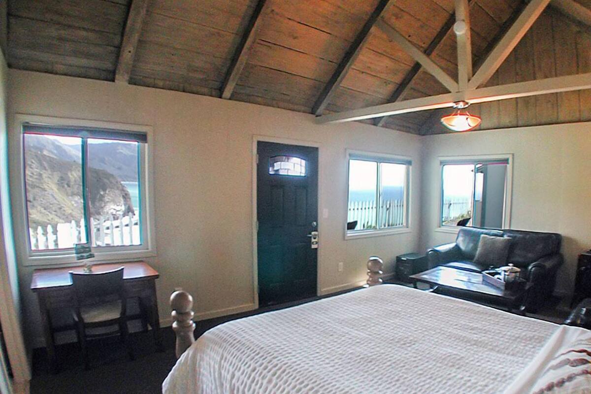 A room with a wooden ceiling, a bed with a white quilt, a desk with a chair, and a sofa.