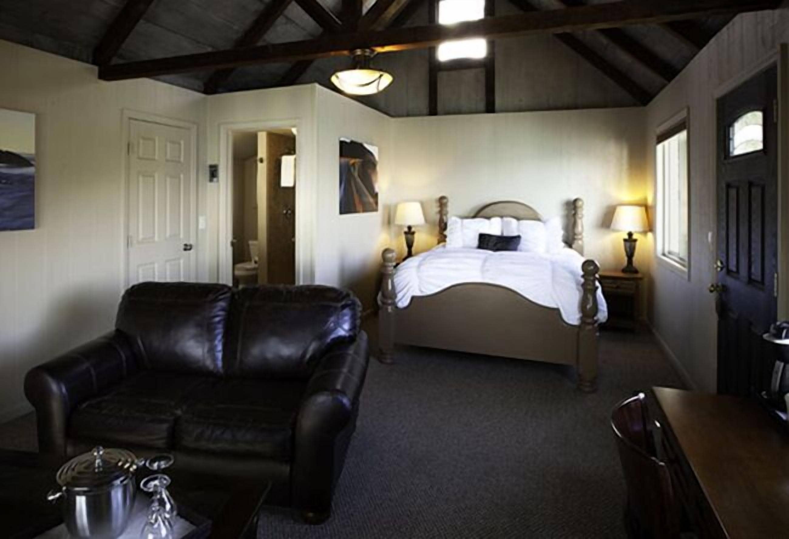 A bedroom with a bed, a black leather chair, a bedside table with a lamp, and a door.