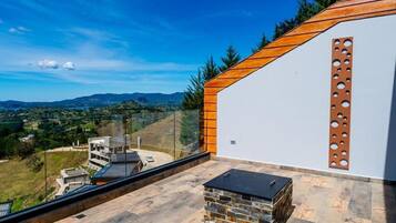 Una terraza moderna al aire libre con una chimenea de piedra, barandilla de cristal y vistas a un paisaje montañoso.
