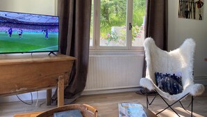 Un salon avec un téléviseur diffusant un match de football, une table en bois, un fauteuil en osier et une fenêtre avec des rideaux.