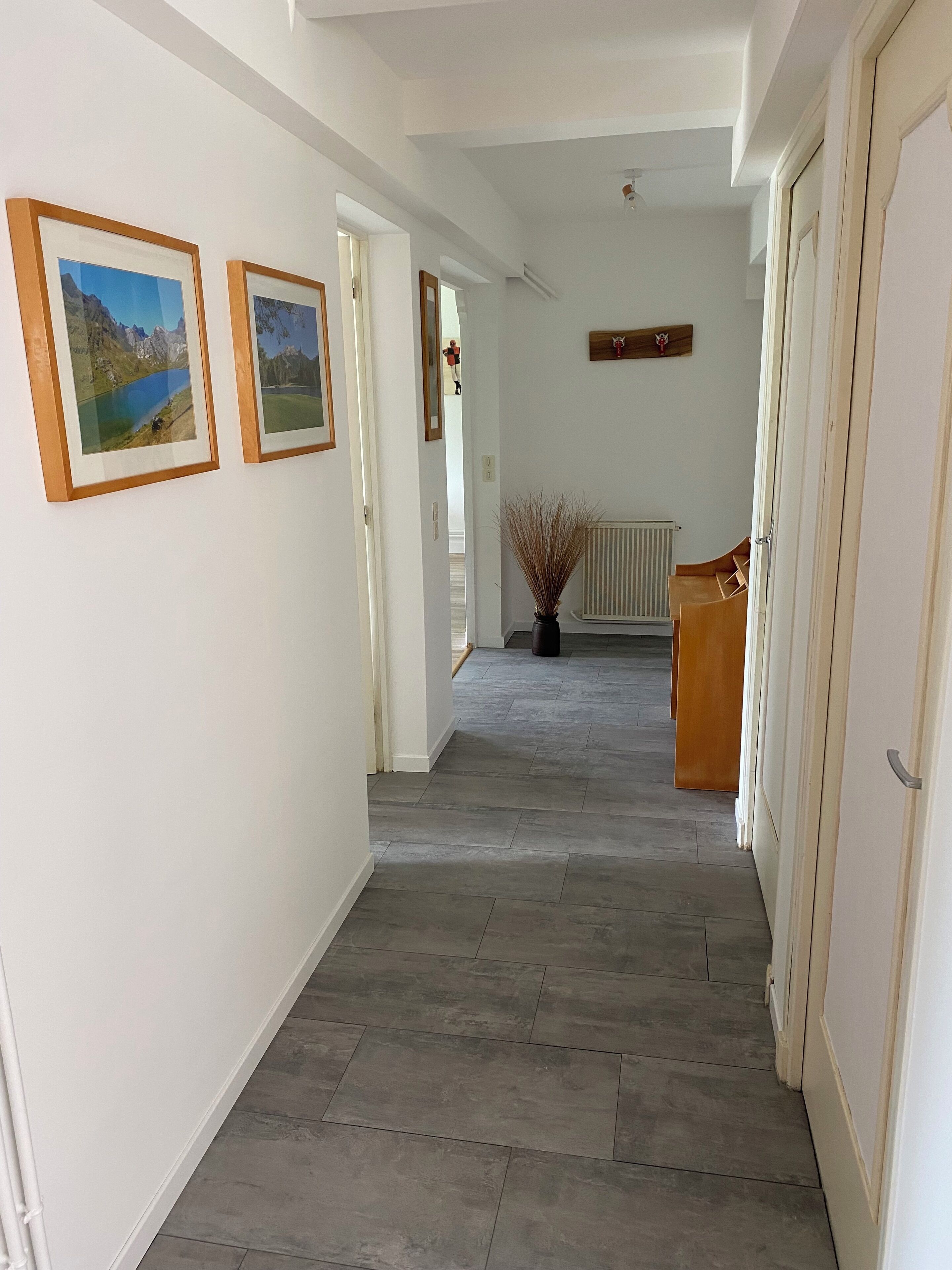 Een hal met ingelijste landschaptaferelen aan de muur, een radiator en een houten deur aan de rechterkant.