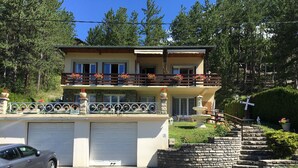 Une maison à deux étages avec un garage, un balcon agrémenté de plantes en pot et un escalier en pierre menant à l’entrée.