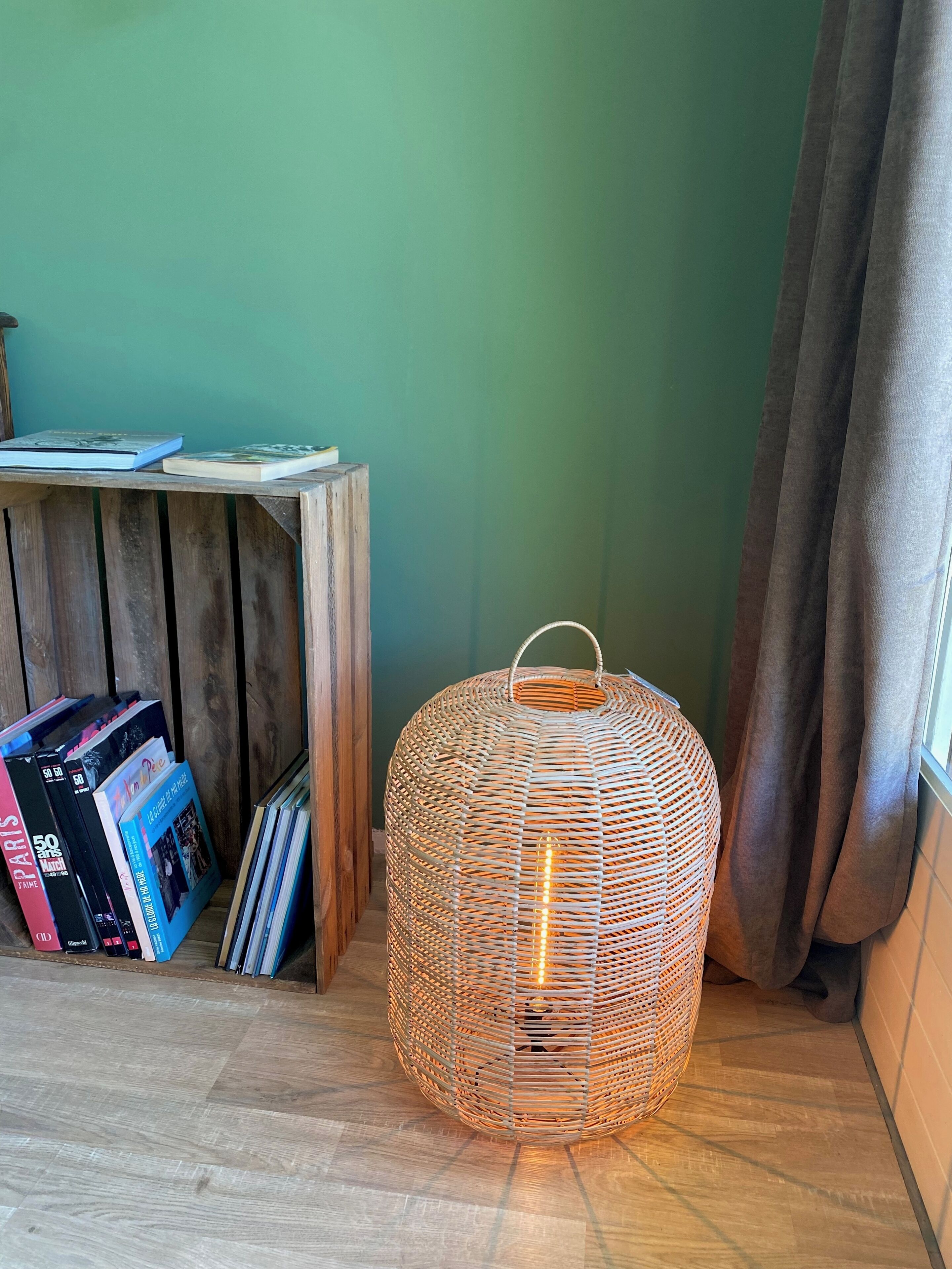 A wicker lamp with a handle, a wooden table with books, and a green wall.
