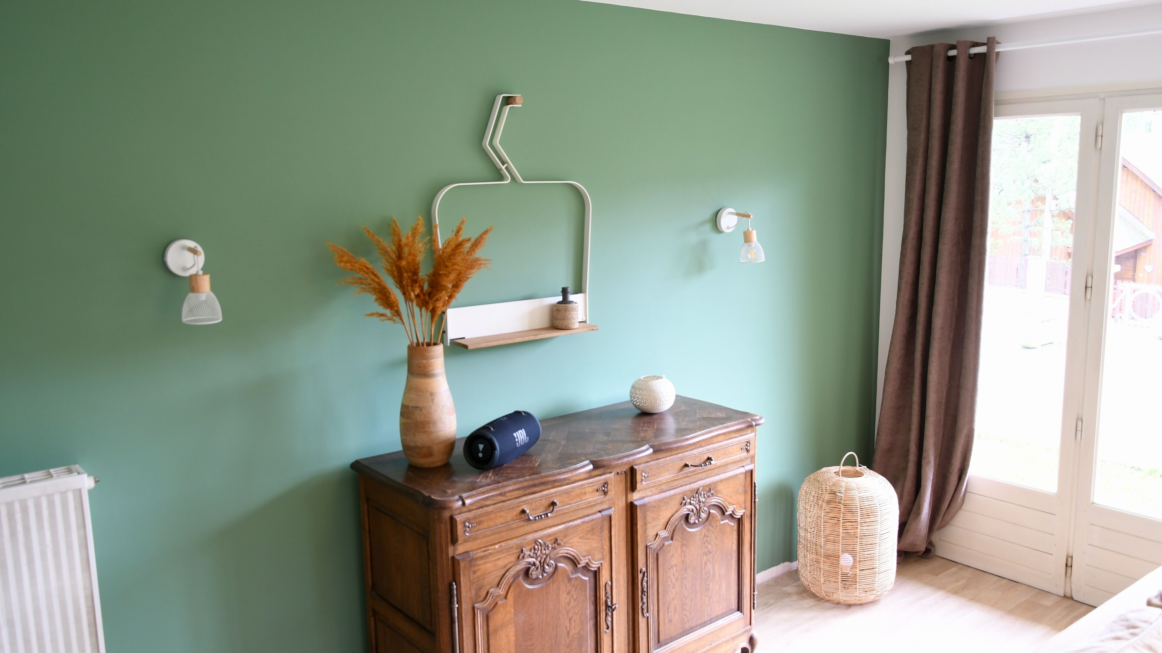 Une pièce avec un mur vert, une commode en bois, un vase contenant des plantes séchées, une petite étagère et une lampe en osier.