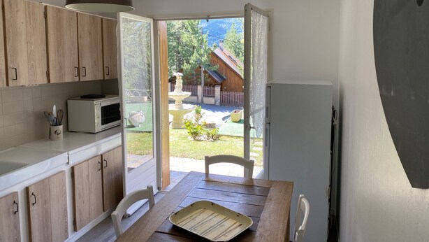 Une cuisine avec des meubles en bois, un four à micro-ondes, une table avec un plateau, et une porte ouverte donnant sur une cour arrière avec une clôture en bois et un abreuvoir à oiseaux.