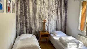 Une chambre avec deux lits simples, une table de chevet en bois, une horloge murale et une fenêtre donnant sur l’extérieur.