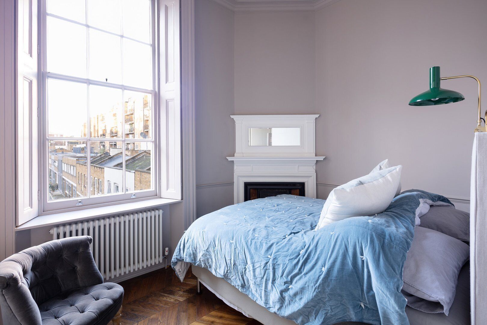 Une chambre avec une grande fenêtre, une cheminée, un lit avec une literie bleue, une chaise et une lampe de bureau verte.