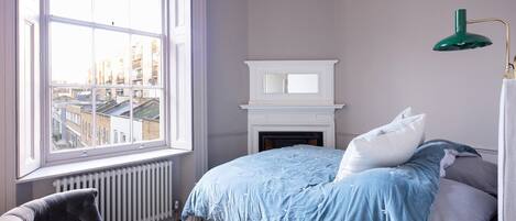 Une chambre avec une grande fenêtre, une cheminée, un lit avec une literie bleue, une chaise et une lampe de bureau verte.