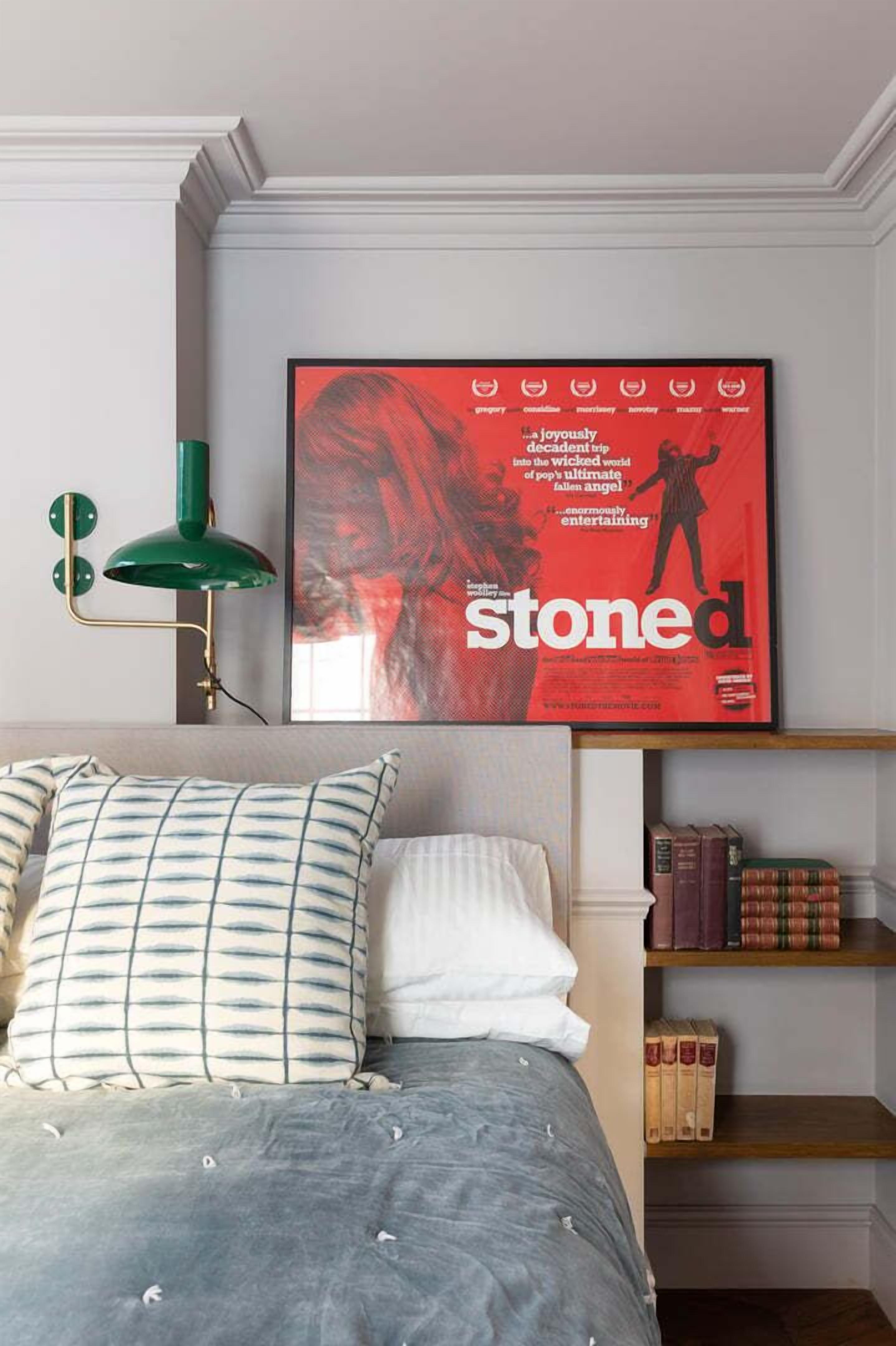 A bedroom with a bed, pillows, a green floor lamp, a framed magazine on the wall, and a bookshelf with books.