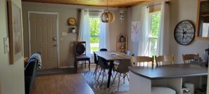 Un espace repas comprenant une table et des chaises en bois, un lustre et une vue sur l’extérieur grâce à de grandes fenêtres.