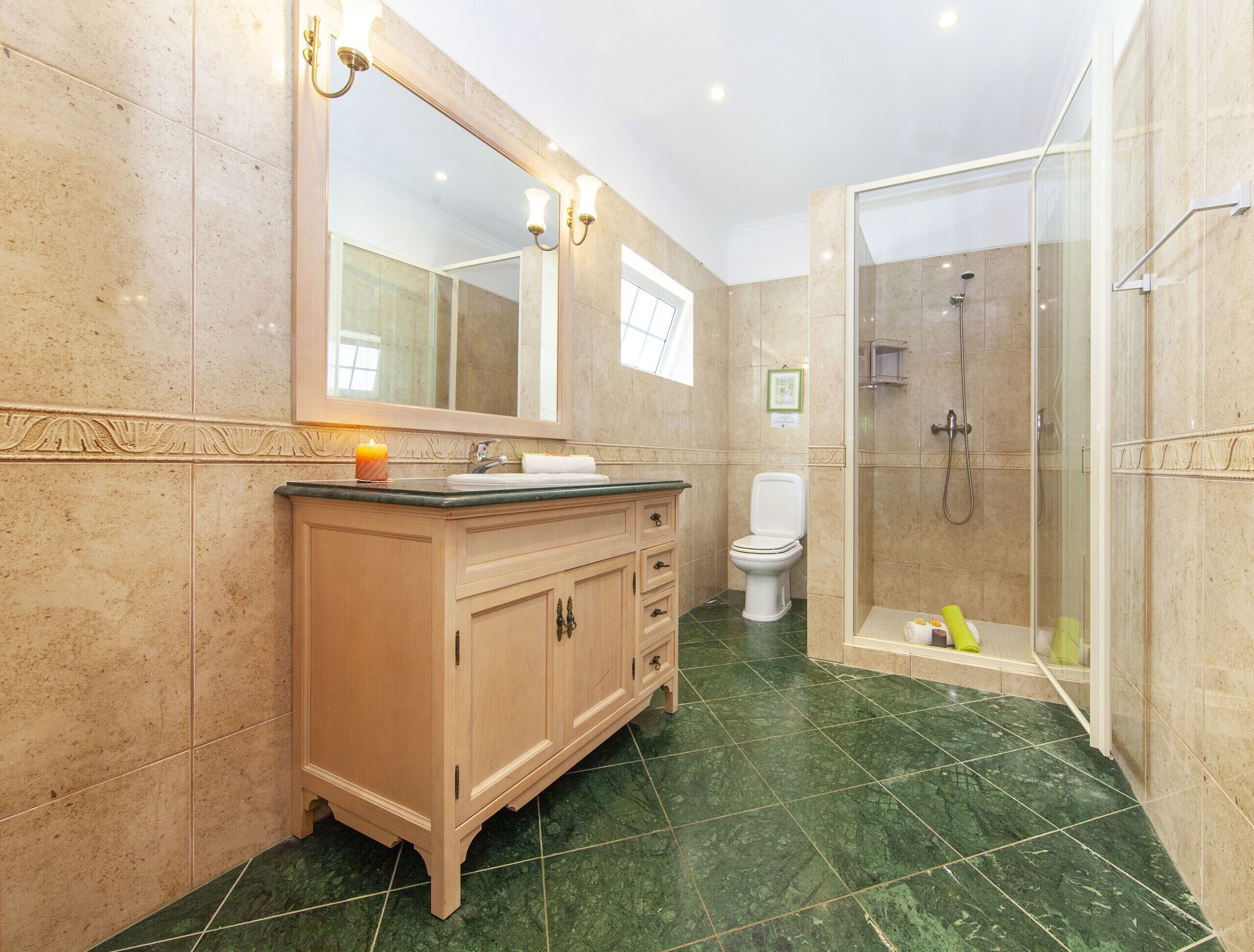 Un baño con piso de mármol, tocador de madera, un espejo grande, inodoro y zona de ducha con mampara de vidrio.