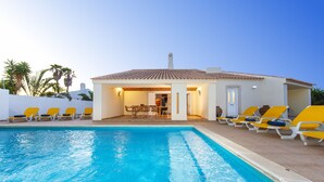 A pool area with a clear blue pool, yellow and white lounge chairs, and a building with a tiled roof. - Villa with heatable pool and BBQ in Albufeira (Albufeira)