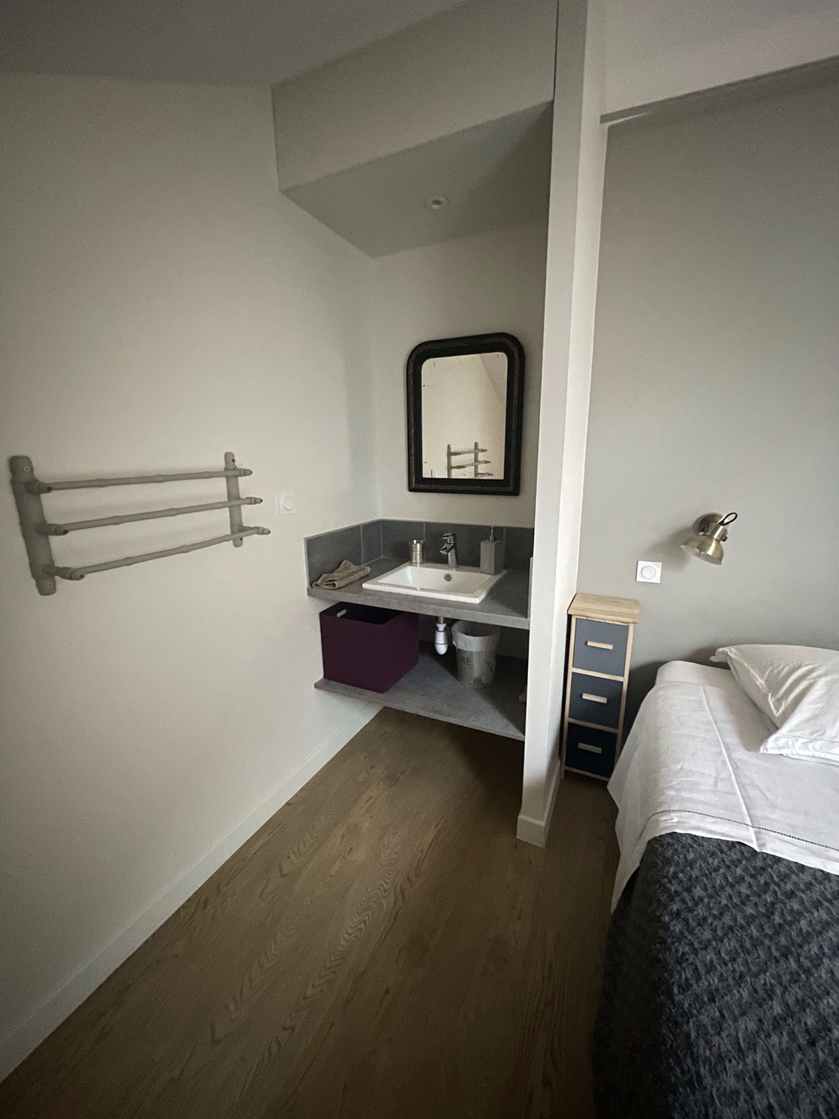 Quarto de hotel compacto com uma cama de solteiro, banheiro pequeno com pia e espelho, e um porta-toalhas.