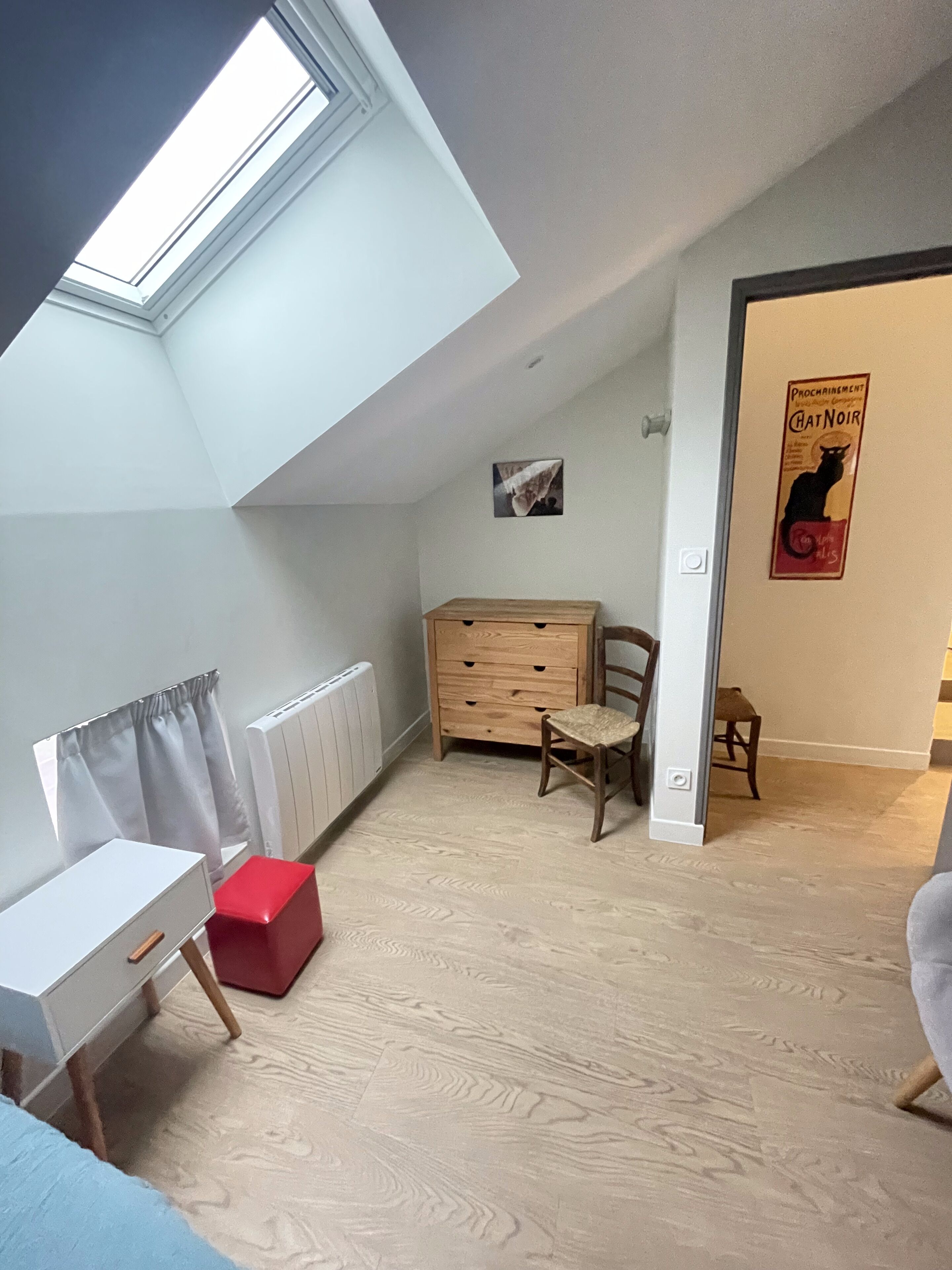 A room with a sloped ceiling, a radiator, a wooden dresser, a small chair, a red storage box, and a poster on the wall.