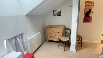 A room with a sloped ceiling, a radiator, a wooden dresser, a small chair, a red storage box, and a poster on the wall.