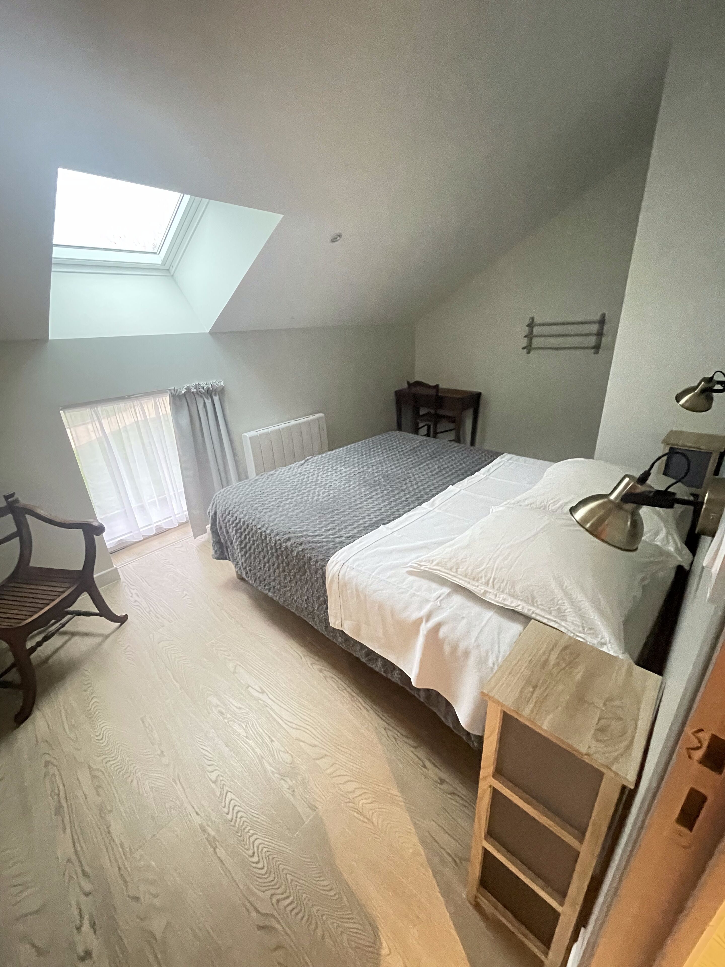 Un dormitorio con tragaluces, una cama con manta blanca y gris, una silla de madera, una mesita pequeña y un radiador.