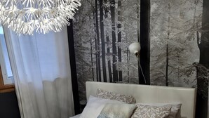 Een netjes opgemaakt bed met een bedspreide met dessin, twee kussens, een muur met een bosdecoratie en een moderne kroonluchter.