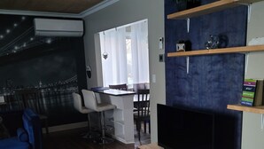 Een moderne woonkamer met een grote televisie, houten vloeren, een blauwe bank, een eethoek met een tafel en stoelen, en een keukenhoek met planken en een magnetron.